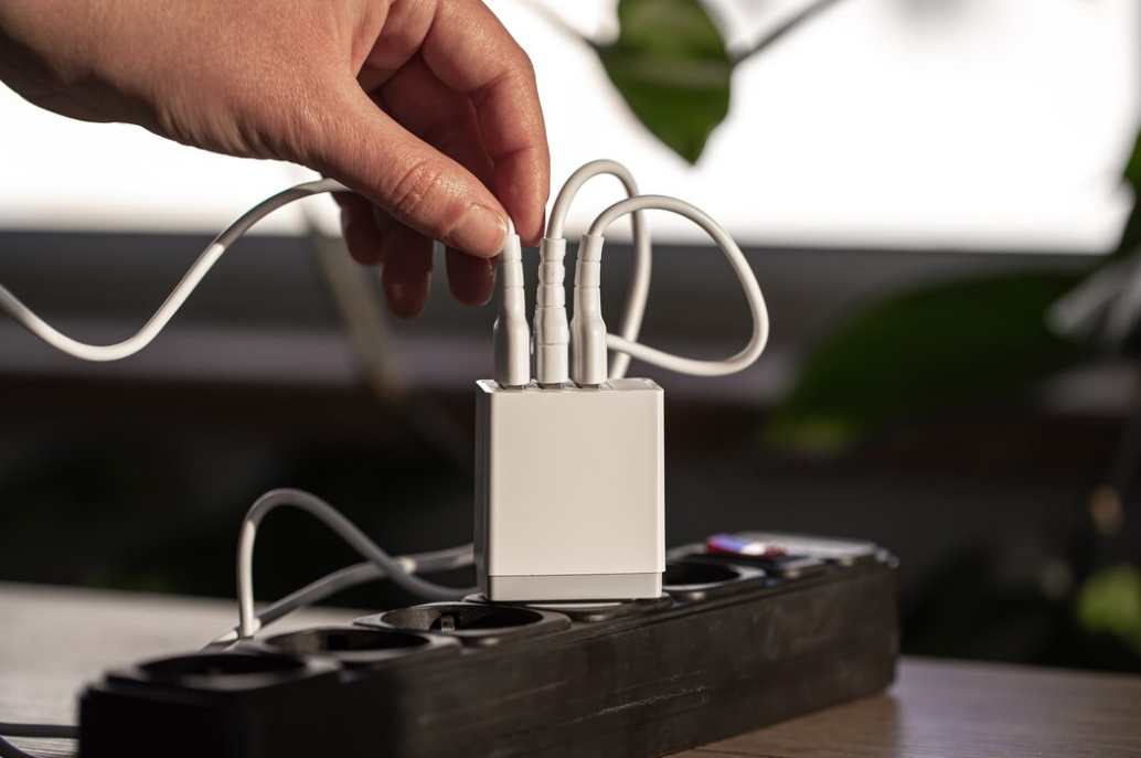 A hand reaching out to adjust cables connected to a white multi-port charging block on a black power strip, with a plant in the background